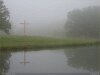 Low contrast photo of a wooden cross on lake shore, reflected in water, foggy